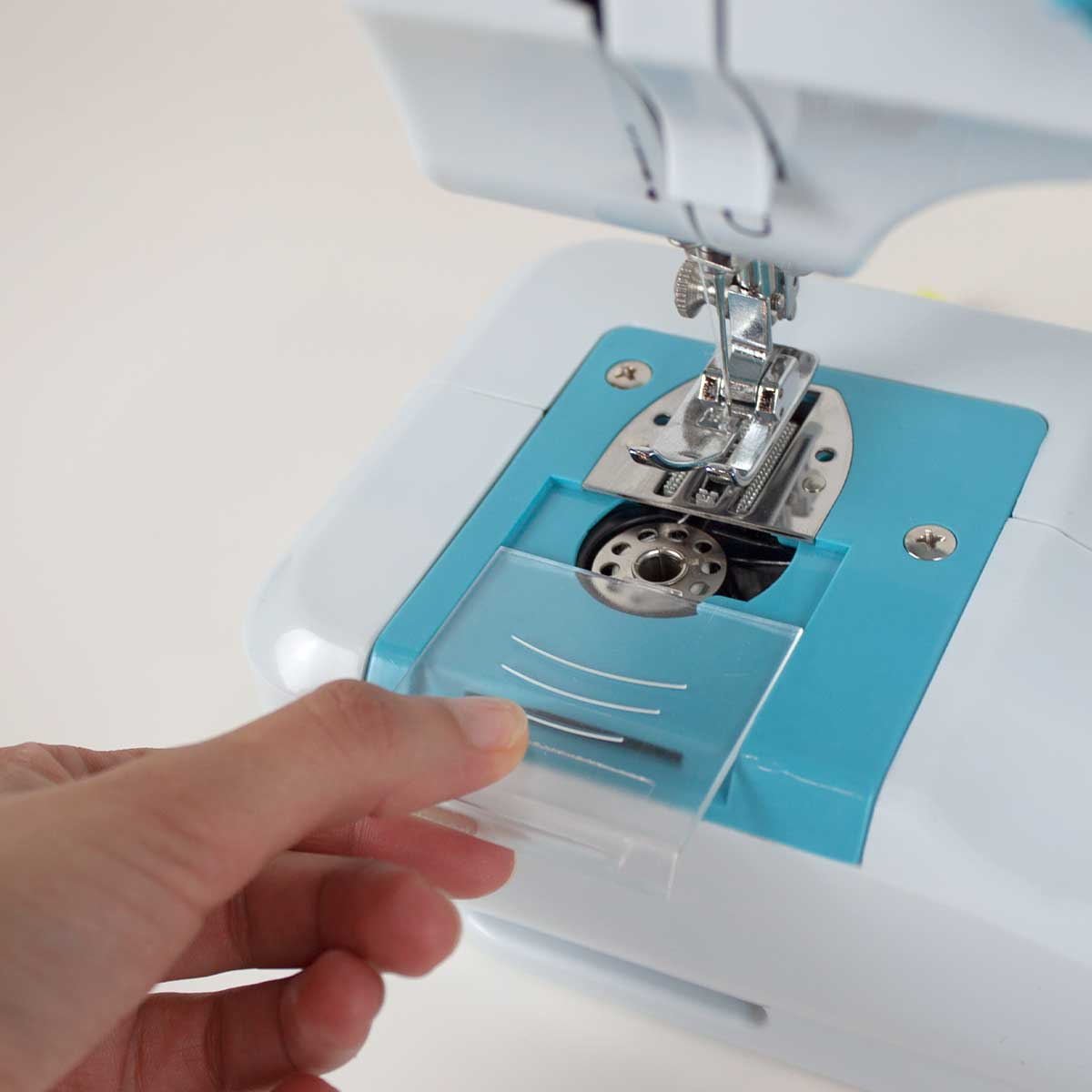 Dłoń otwierająca przezroczystą plastikową osłonę na maszynie do szycia. Maszyna jest biało-niebieska.