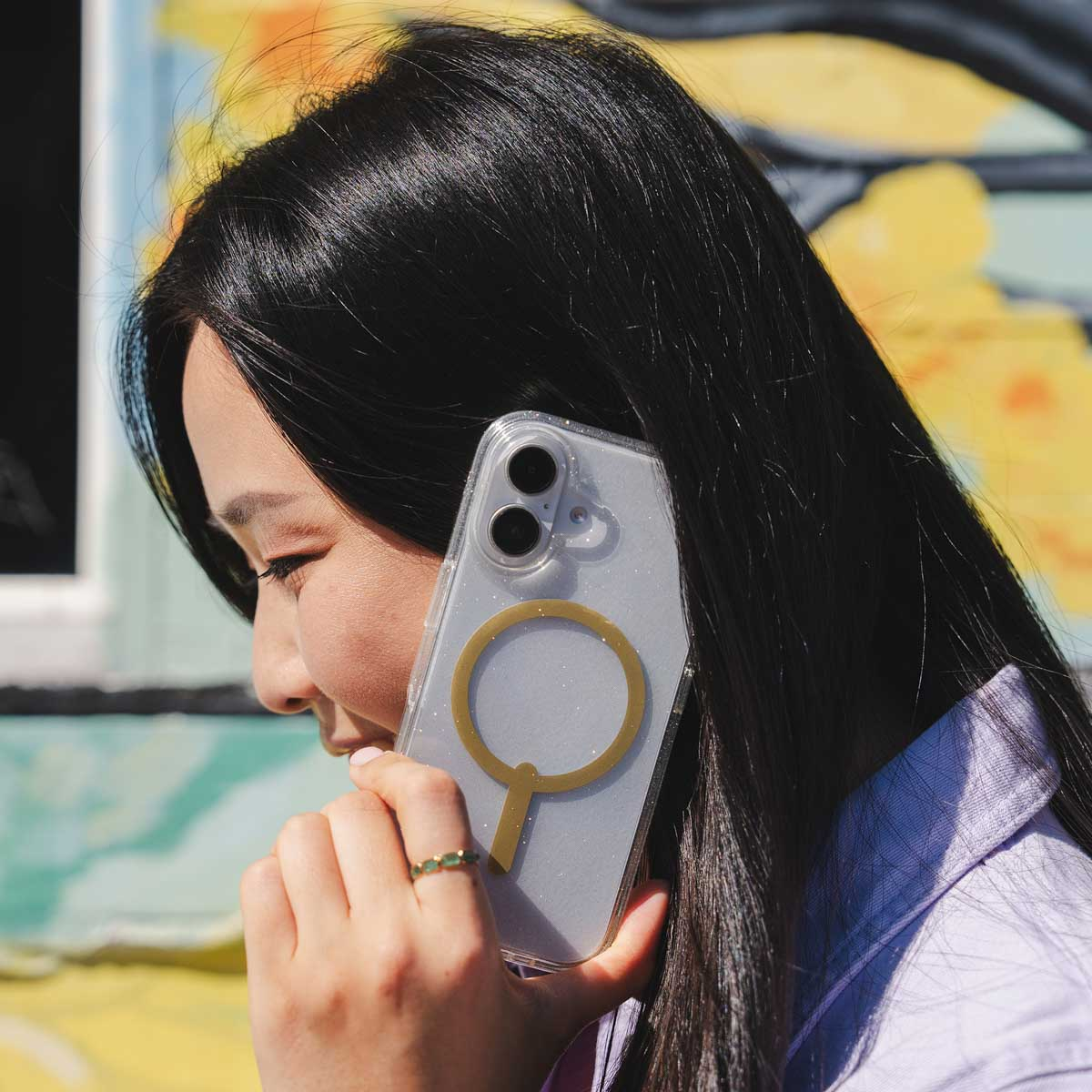 Frau hält ein Telefon in einer durchsichtigen Hülle mit goldenem Ring. Der Hintergrund hat ein Wandgemälde.