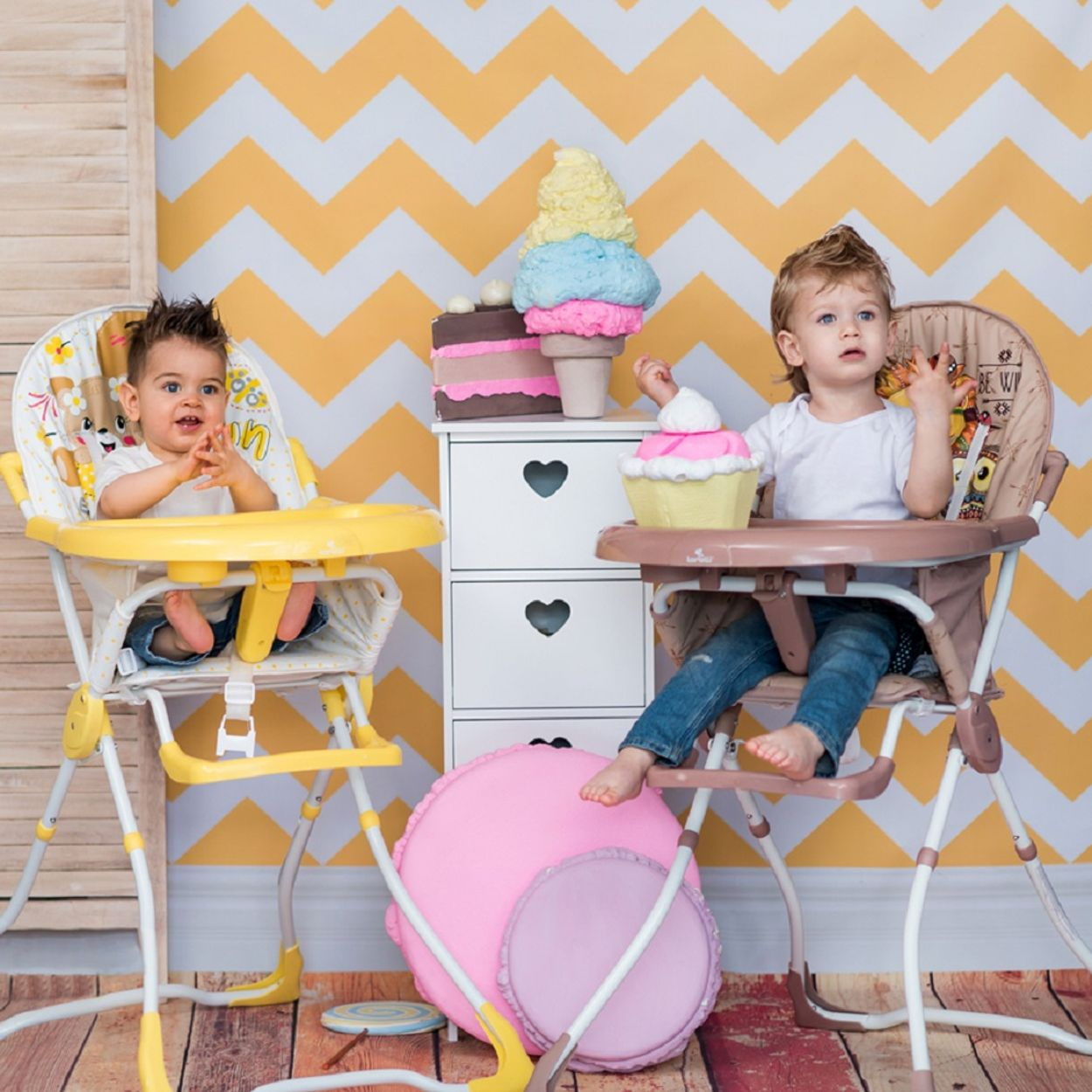 Zwei Babys in Hochstühlen auf einer Geburtstagsfeier. Dekorationen, Kuchen und Spielzeug sind im Hintergrund sichtbar.