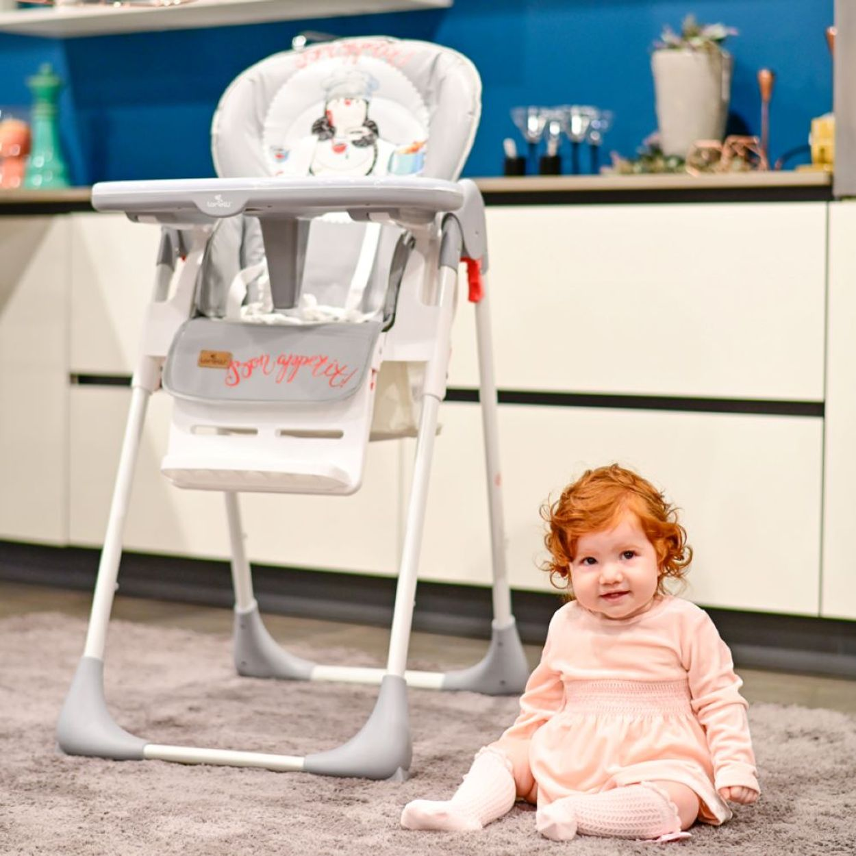 Ein Baby sitzt auf einem grauen Teppich und lächelt. Ein Hochstuhl steht dahinter.