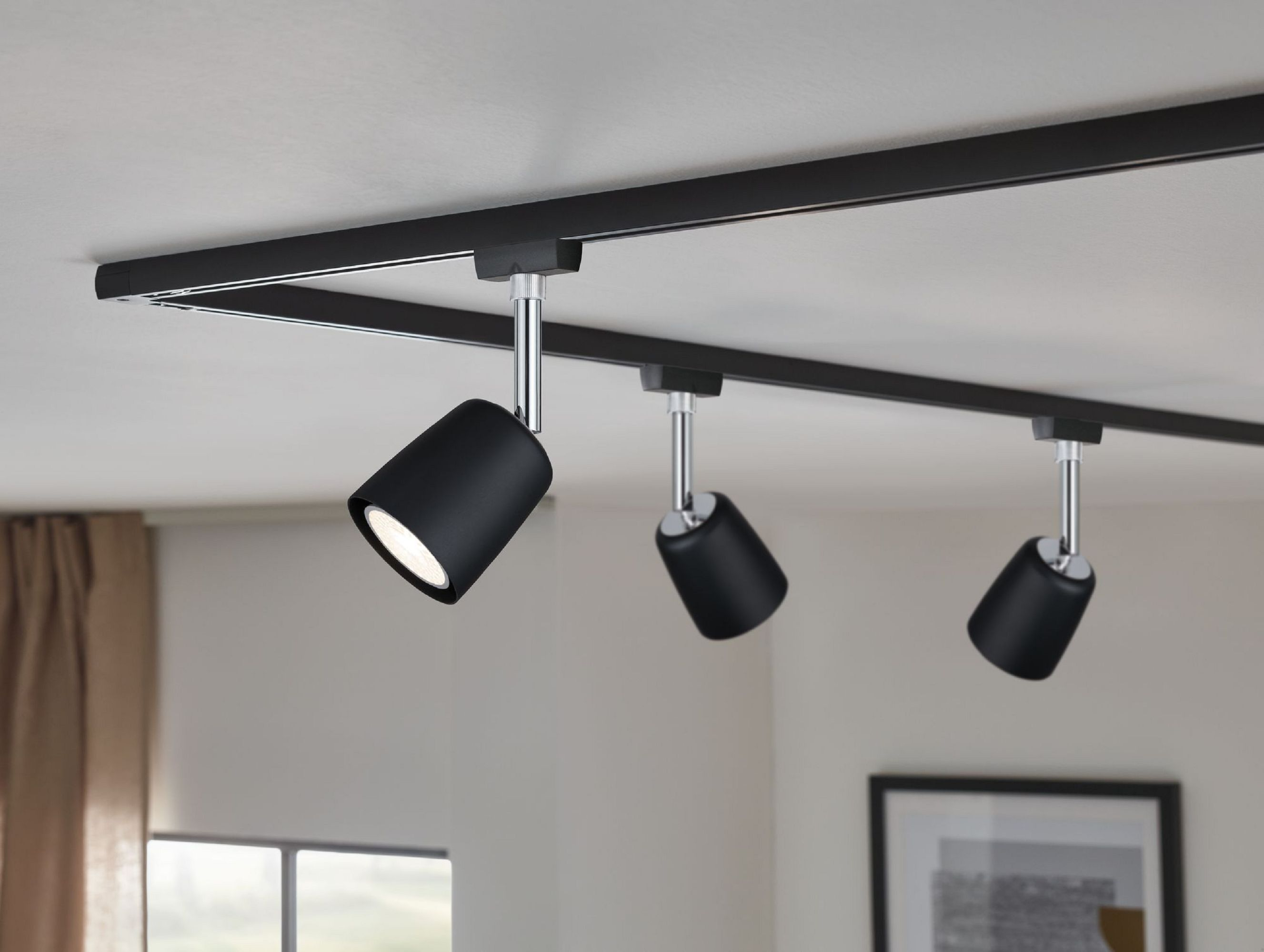 Drei schwarze Strahler an einer schwarzen Schiene, an einer weißen Decke befestigt, mit Wand.