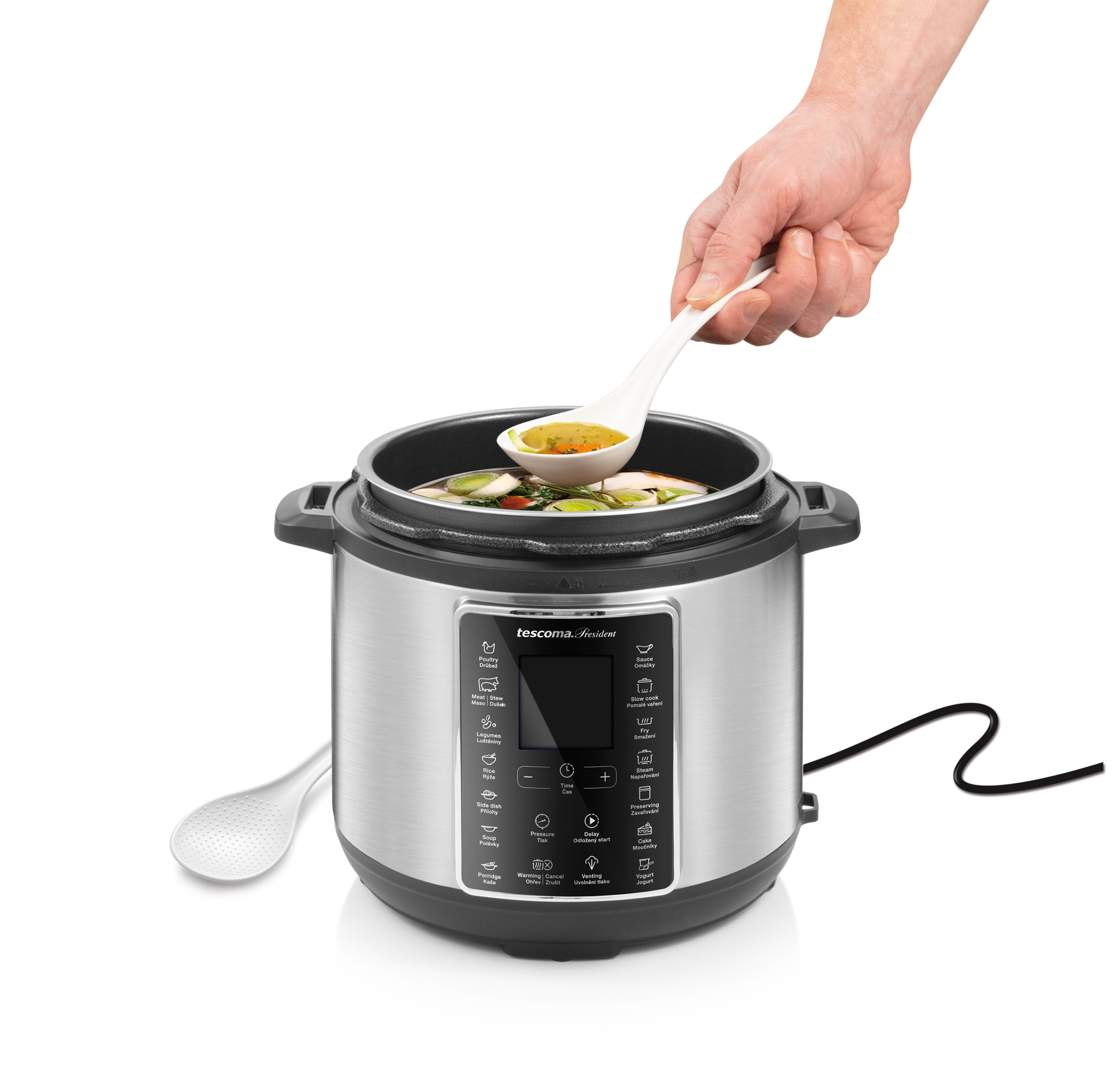 Eine Hand schöpft mit einem Löffel Suppe aus einem silbernen Schnellkochtopf. Ein Löffel liegt daneben.