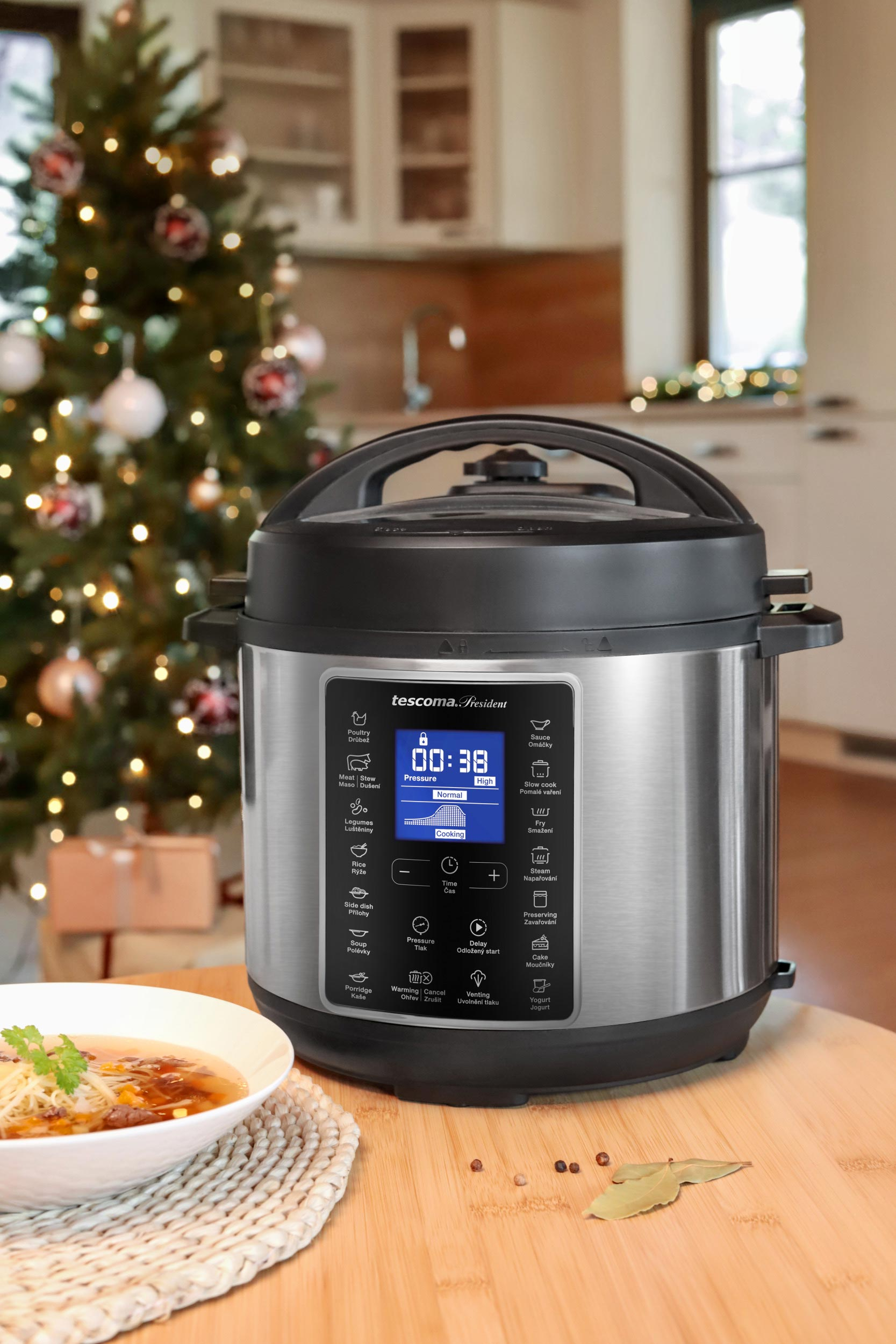 Ein silberner Schnellkochtopf auf einem Holztisch mit Essen, einer weißen Schüssel mit Suppe und einem Weihnachtsbaum im Hintergrund.