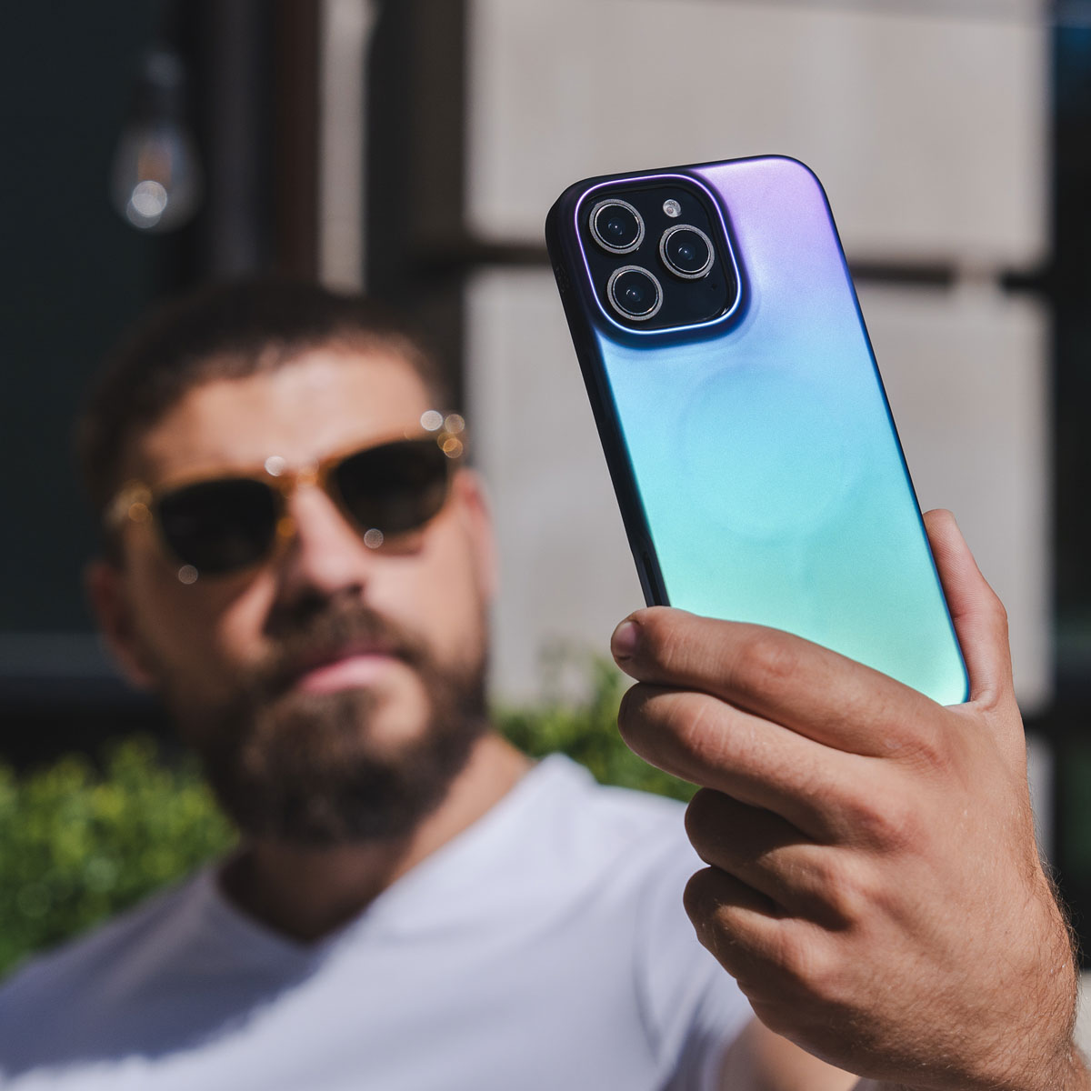 Un homme tient un téléphone, prenant un selfie. La coque a des couleurs irisées. Arrière-plan extérieur flou.