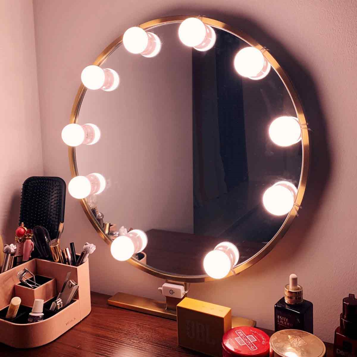 Ein runder Schminkspiegel mit Lichtern, umgeben von Make-up-Zubehör auf einem Tisch.