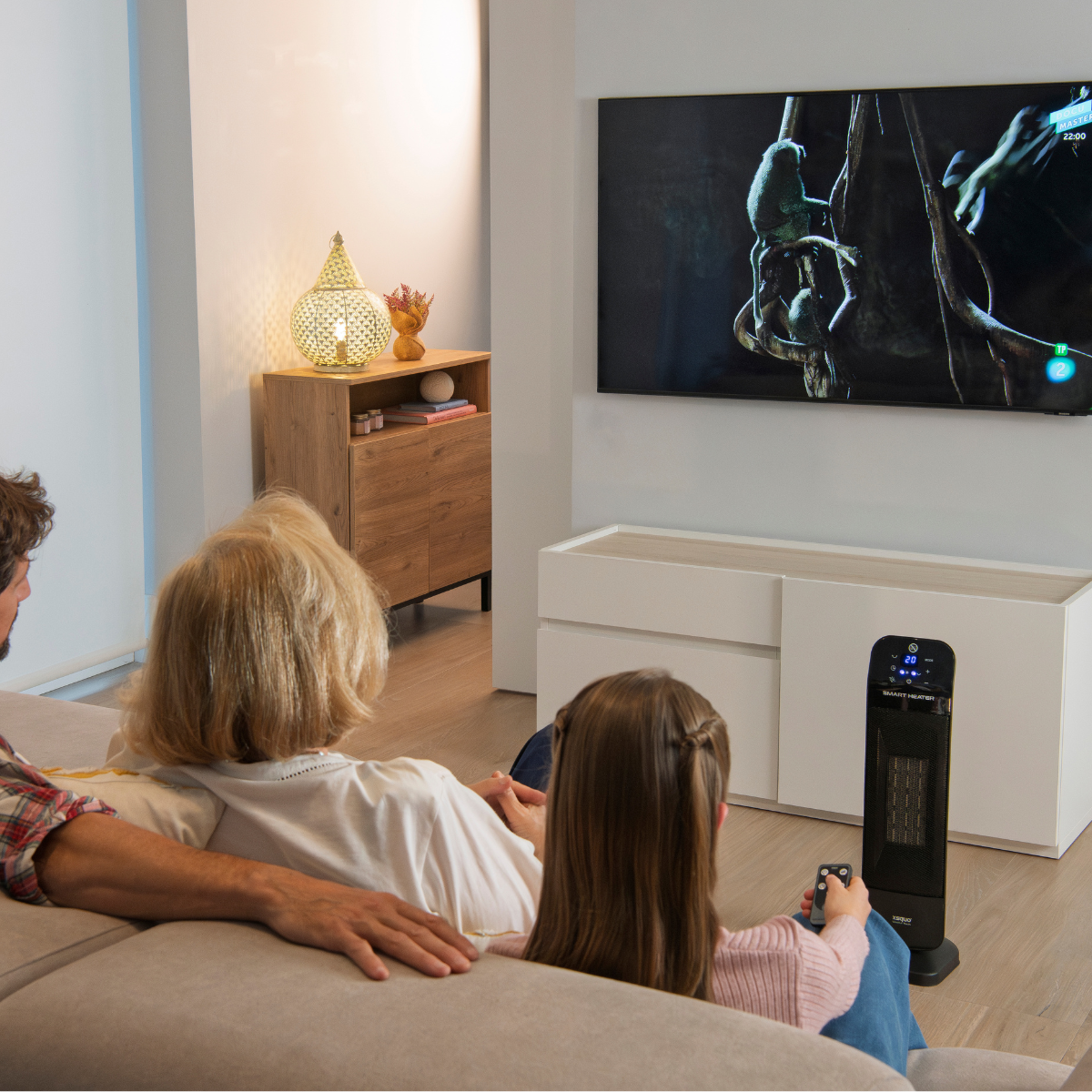 Familia viendo la televisión. Un calentador negro al frente. Mueble de madera y lámpara atrás.
