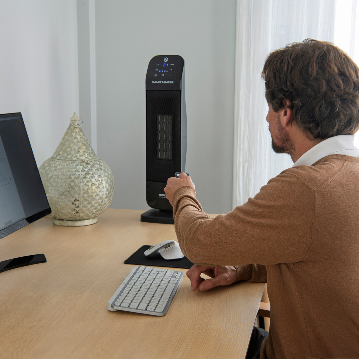 Hombre usando mando a distancia para un calefactor inteligente negro en un escritorio con computadora y teclado.