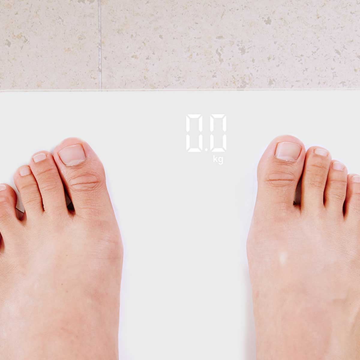 Bose stopy na białej wadze cyfrowej pokazującej '0,0 kg'. Tło: podłoga z płytek.