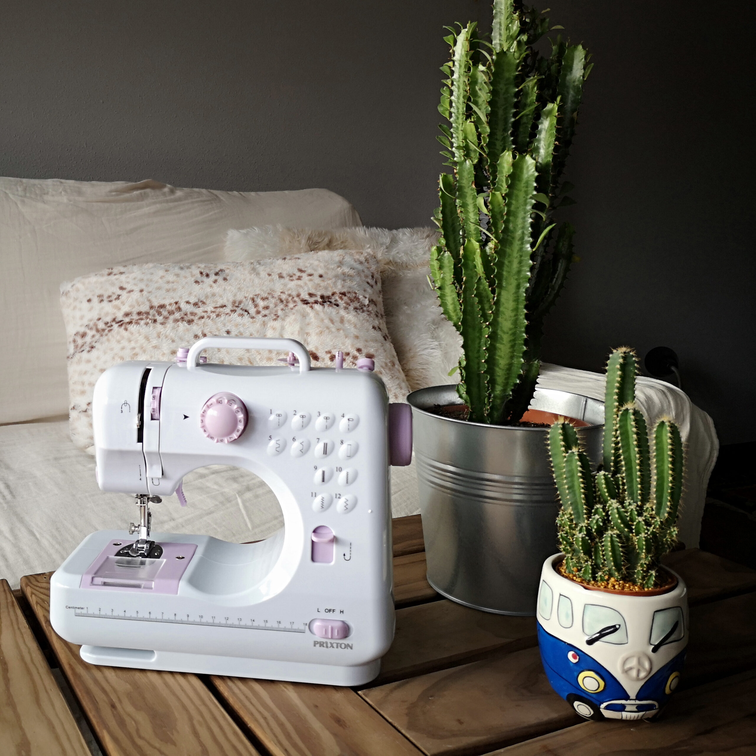Biała maszyna do szycia z różowo-fioletowym designem, kaktusy i poduszki na drewnianym stole.