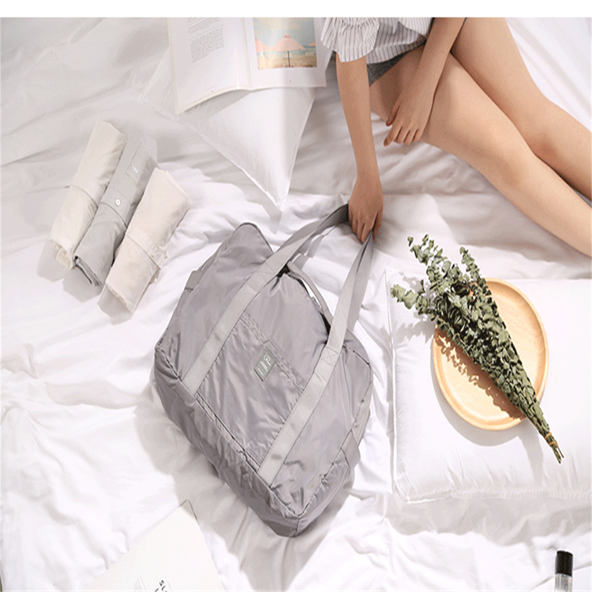 Eine Hand einer Frau hält eine graue Tasche auf einem Bett. Gefaltete Hemden und Blumen liegen auf dem Bett.