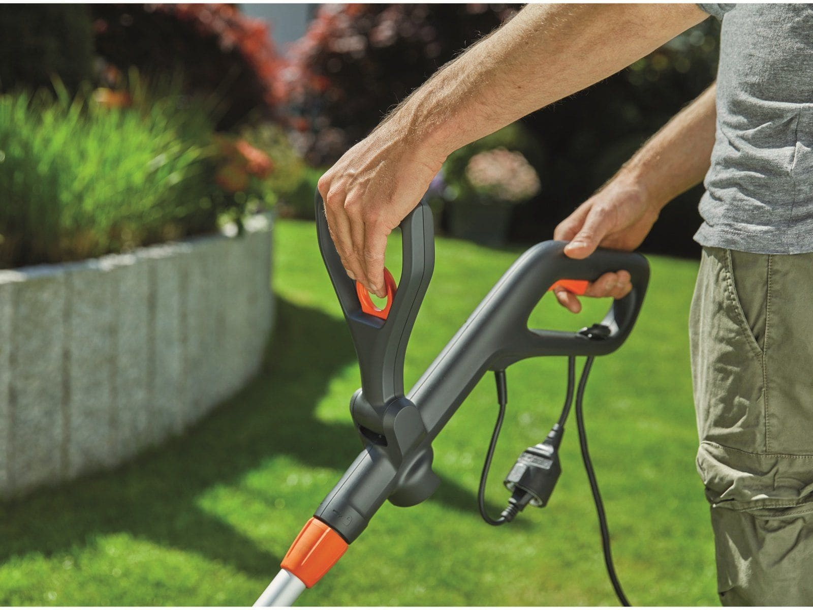 Person hält einen schwarz-orangen Trimmergriff in einem grasbewachsenen Garten, mit einem Netzkabel.