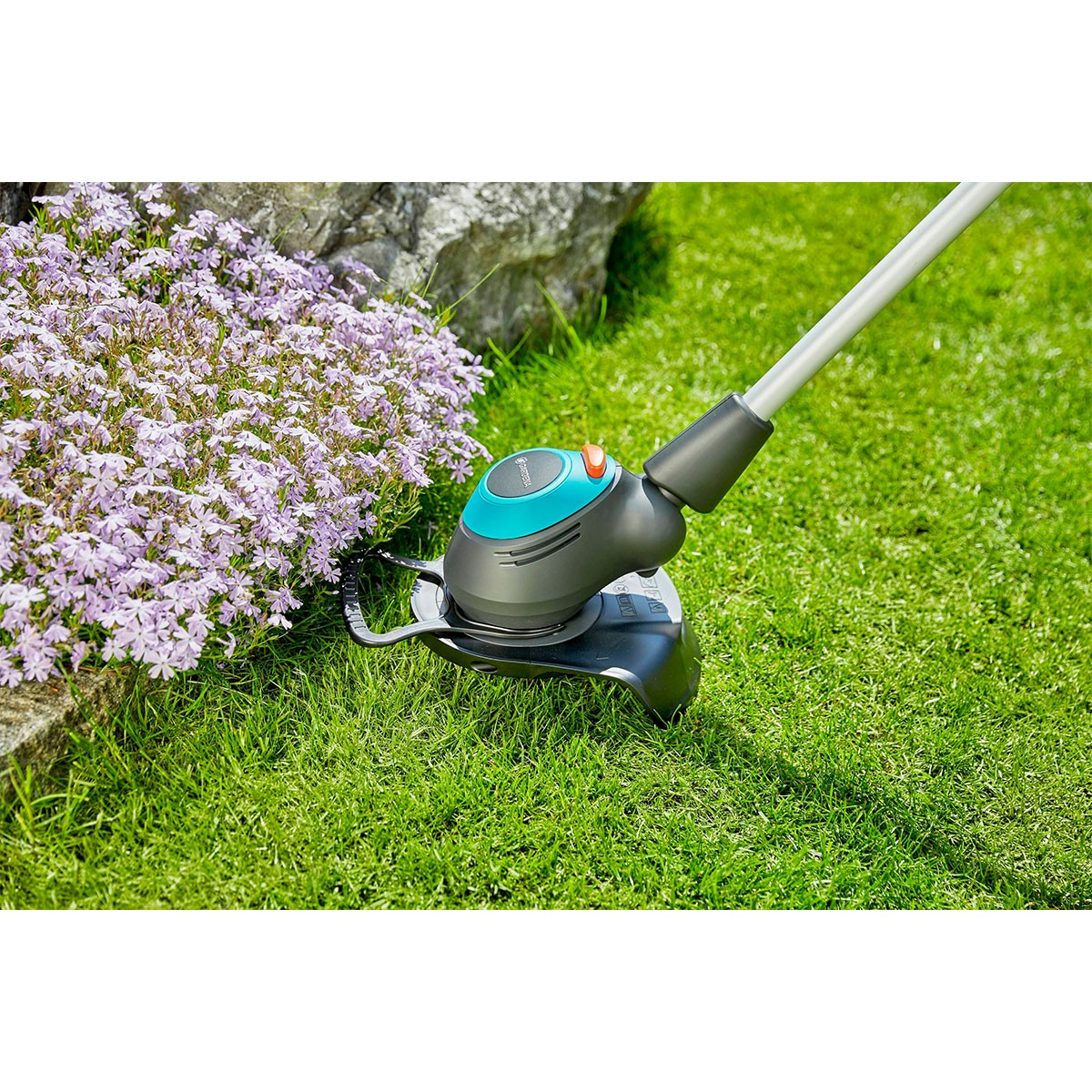 Ein grüner Rasenmäher schneidet Gras in der Nähe eines lila Blumenbeets.