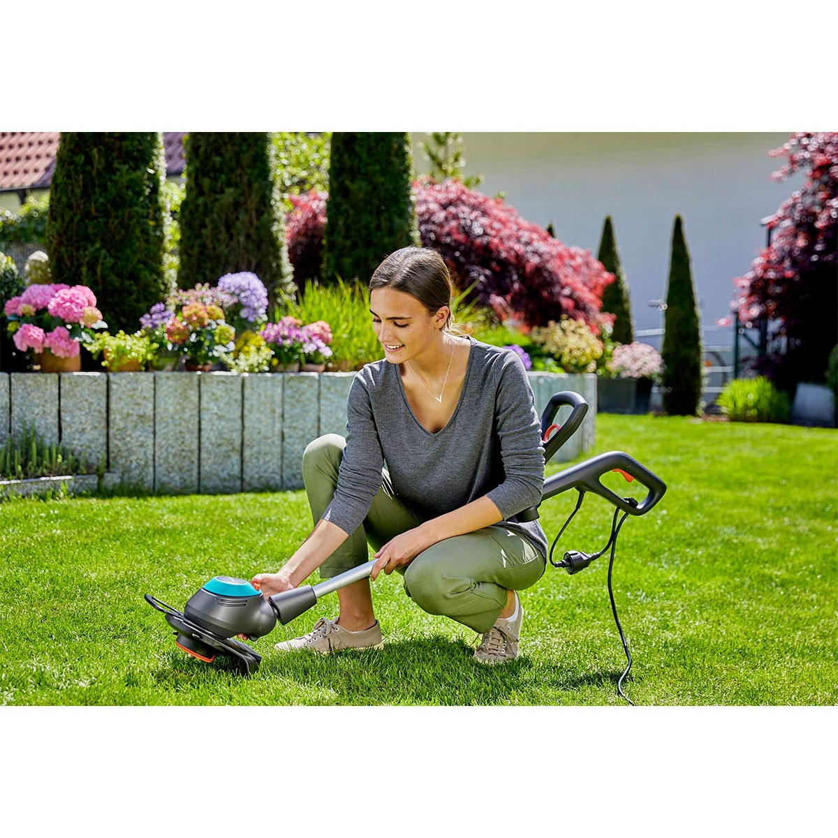 Frau benutzt einen Gardena-Grasschneider in einem Garten, trimmt Gras in der Nähe von Blumen und Büschen.