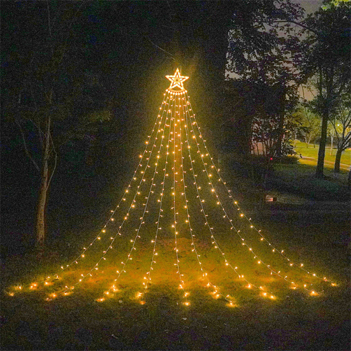 Nachts: ein leuchtender Weihnachtsbaum aus Lichtern in einem Park mit Bäumen. Oben ist ein Stern.