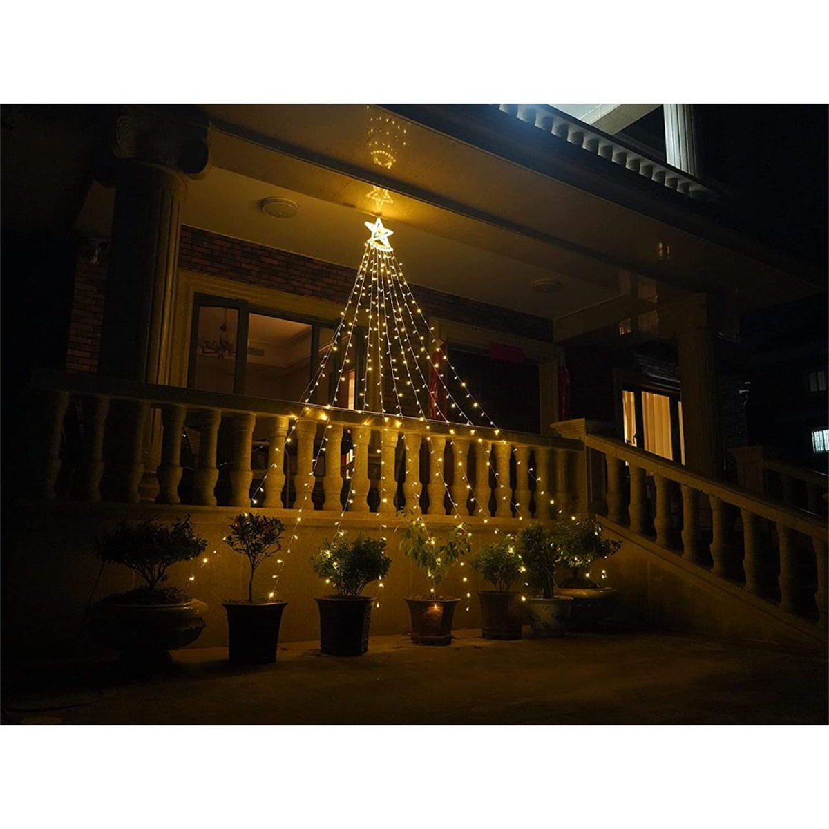 Weihnachtsbaumlichter hängen an einem Haus. Stern oben. Pflanzen und ein Zaun. Nachtszene.