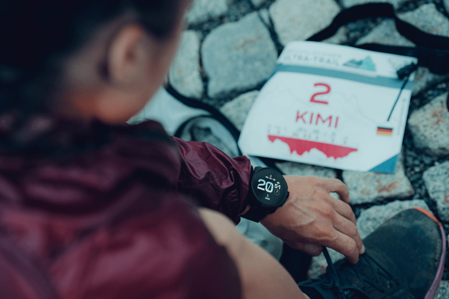 Ręka osoby ze smartwatchem. Zawiązuje sznurowadło. Numer startowy w tle, z tekstem i flagą.