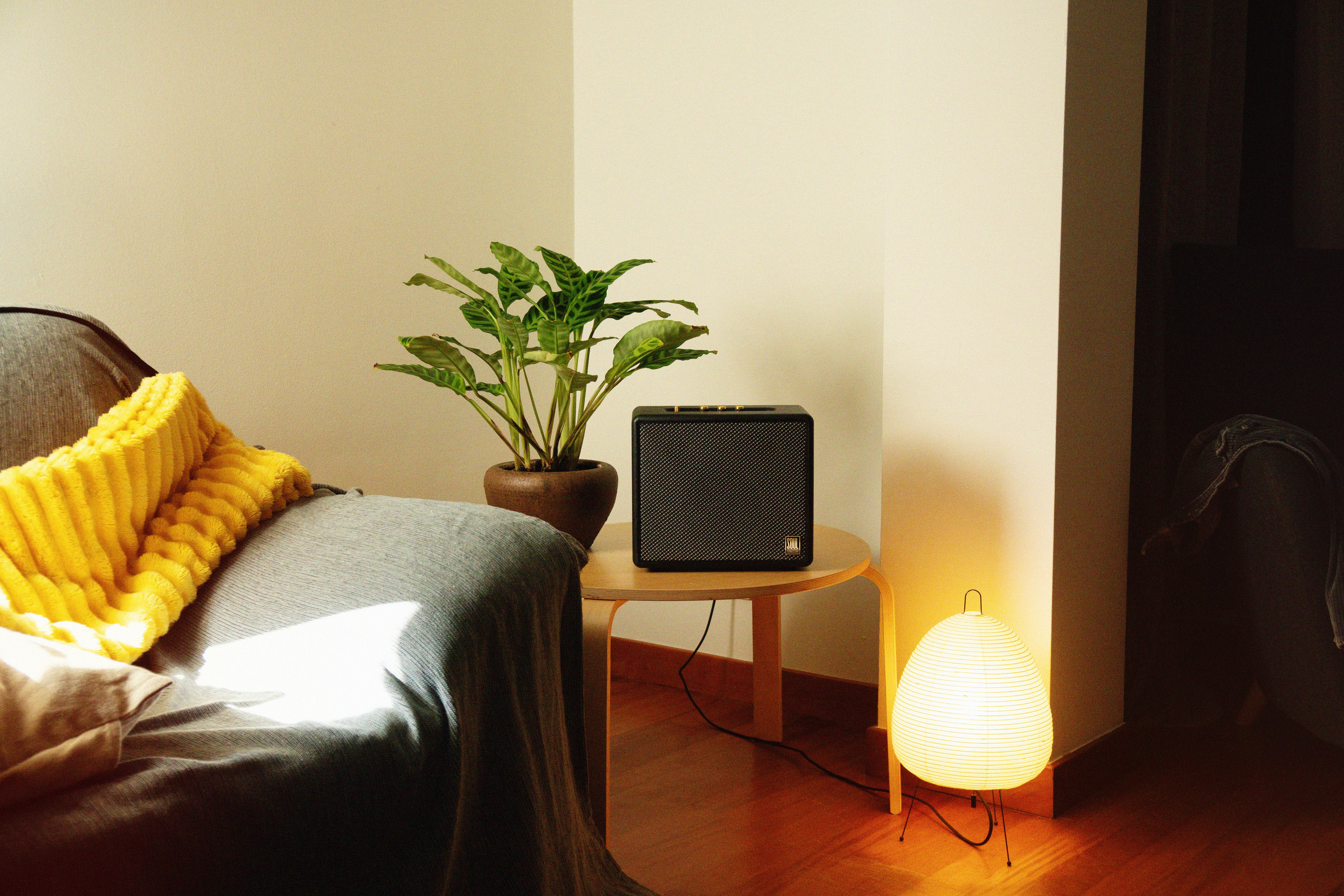 Un altavoz negro sobre una mesa, con un sofá y una planta al fondo, y una lámpara.