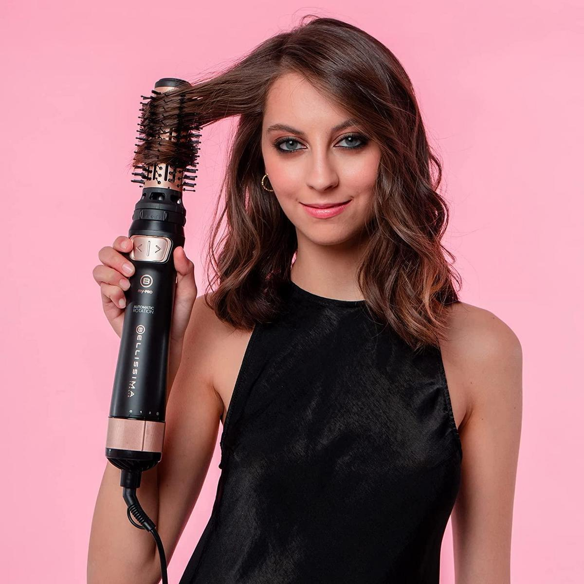 Mujer peinando su cabello con un moldeador de cabello negro y dorado, fondo rosa.