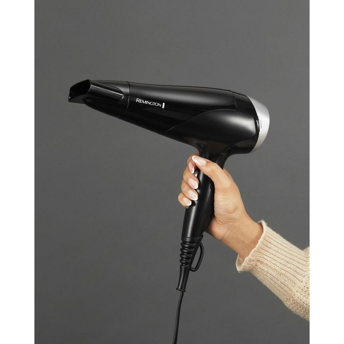 De hand van een persoon die een zwarte haardroger vasthoudt. De haardroger is zilver en zwart, met het merk Remington.