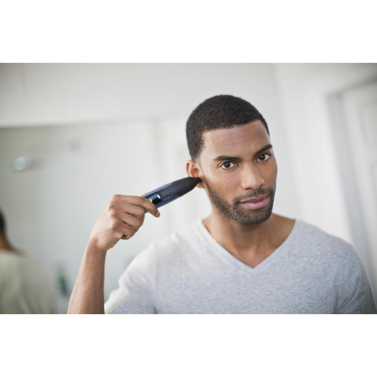 Mężczyzna używa czarnej trymerki do brody przed lustrem. Ma na sobie szarą koszulę.