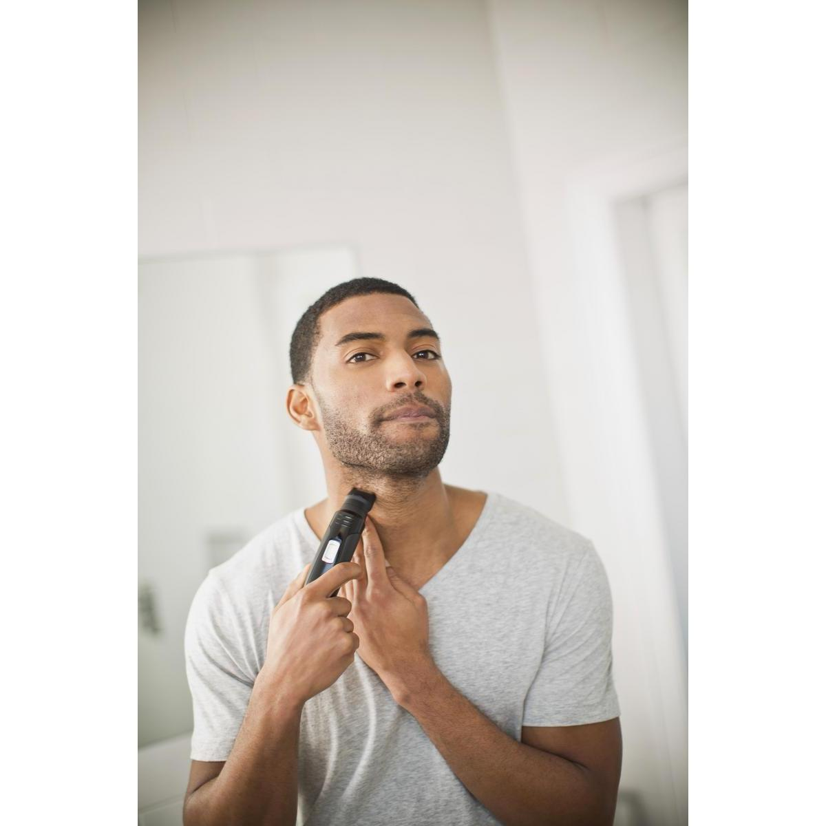 Mężczyzna goli szyję elektryczną maszynką do golenia przed lustrem. Ma na sobie szarą koszulę.