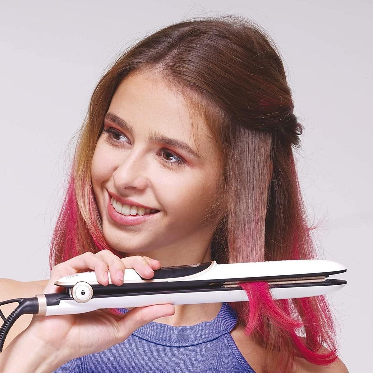 Eine Frau benutzt einen Haarglätter auf pink gefärbtem Haar. Der Glätter ist weiß, die Frau lächelt.