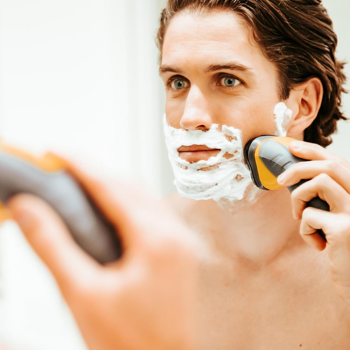 Un uomo si rade la barba davanti a uno specchio, usando un rasoio elettrico.