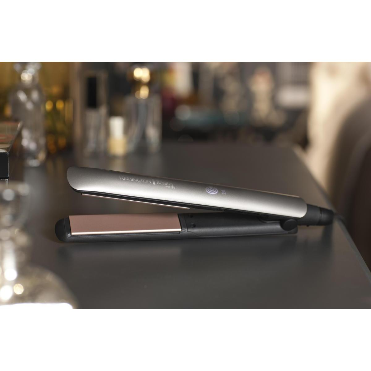 Srebrna prostownica do włosów z płytką w kolorze różowego złota. Leży na szarej powierzchni.