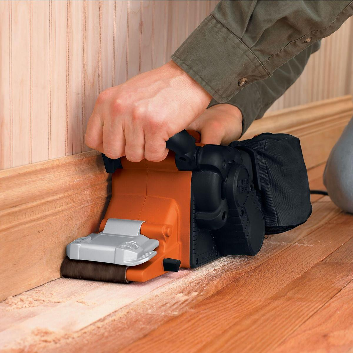 Person schleift Holzfußleiste mit orangem Schleifer, Sägemehl sichtbar.
