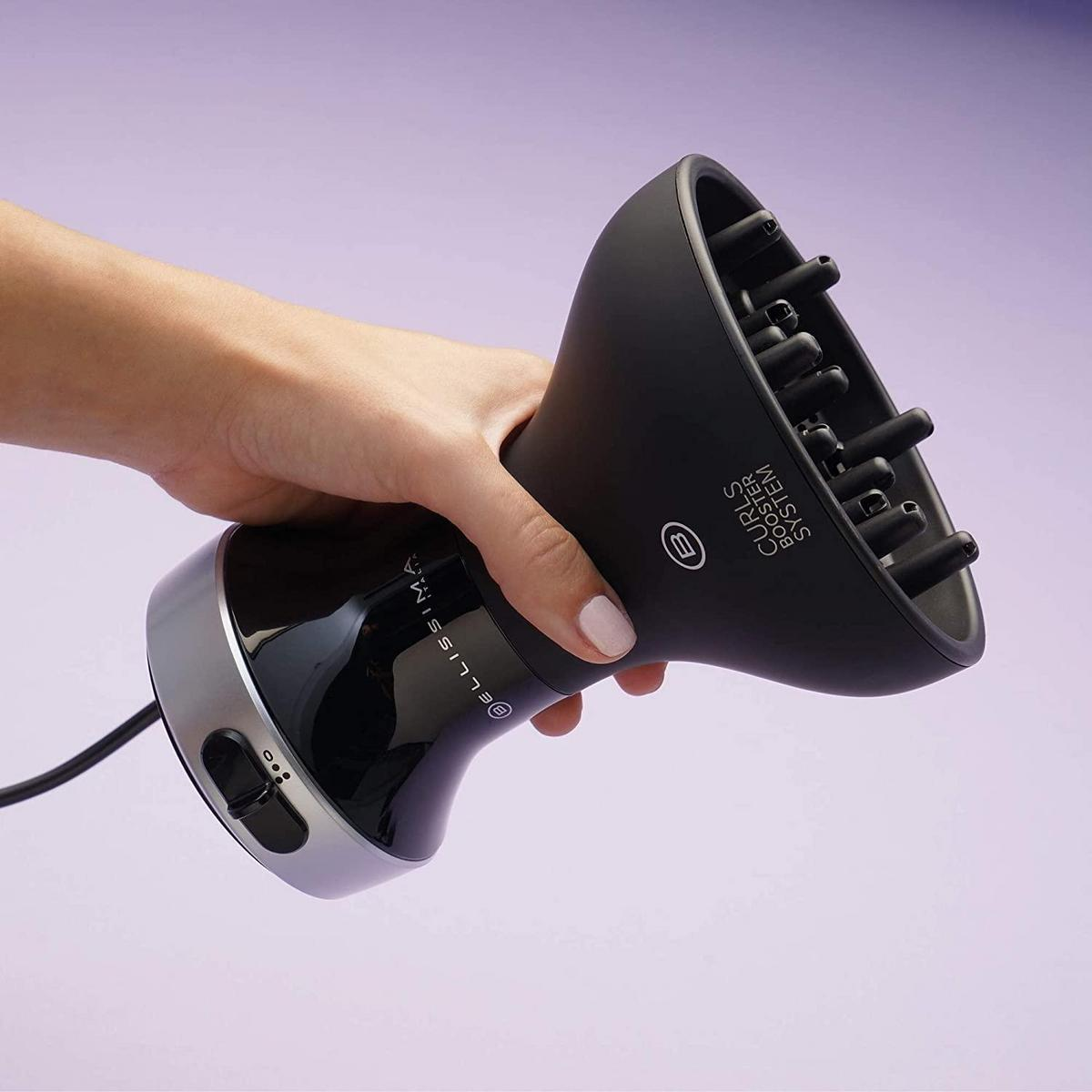 Una persona tiene in mano un diffusore per capelli nero con base argentata su sfondo viola.