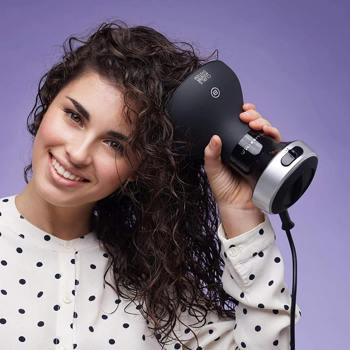 Donna con capelli ricci usa un asciugacapelli nero, sorridente, con sfondo viola.