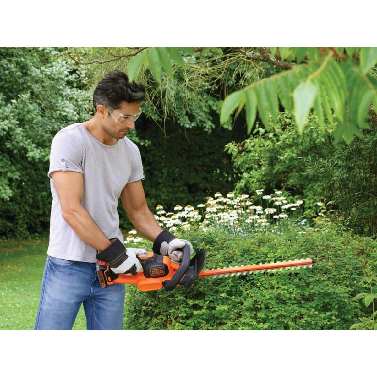 Mann mit Schutzbrille und Handschuhen, der eine Hecke mit einer orangefarbenen BLACK+DECKER beschneidet.
