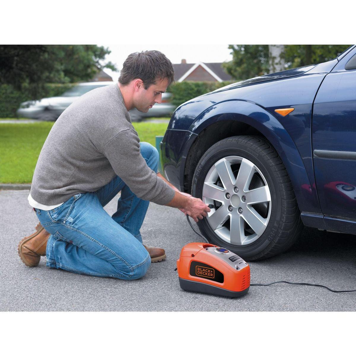 Ein Mann kniet und pumpt einen blauen Autoreifen mit einem orangefarbenen Black+Decker-Inflator auf einer gepflasterten Oberfläche auf.