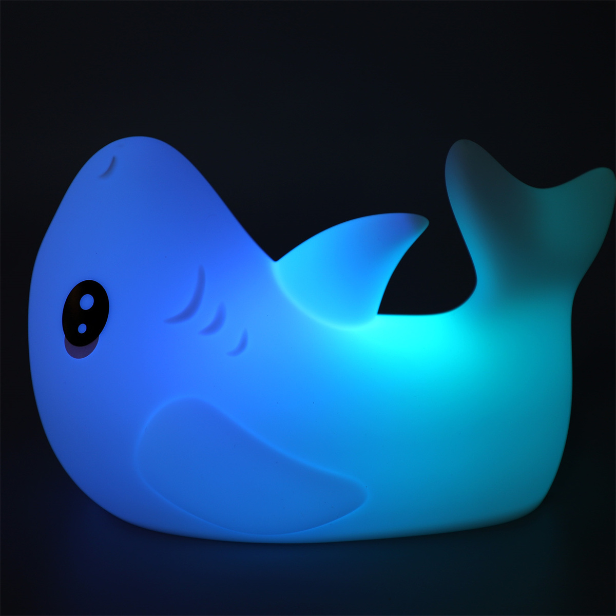 Ein blaues, haiförmiges Nachtlicht leuchtet auf dunkler Oberfläche. Es hat ein fröhliches Gesicht und Flossen.