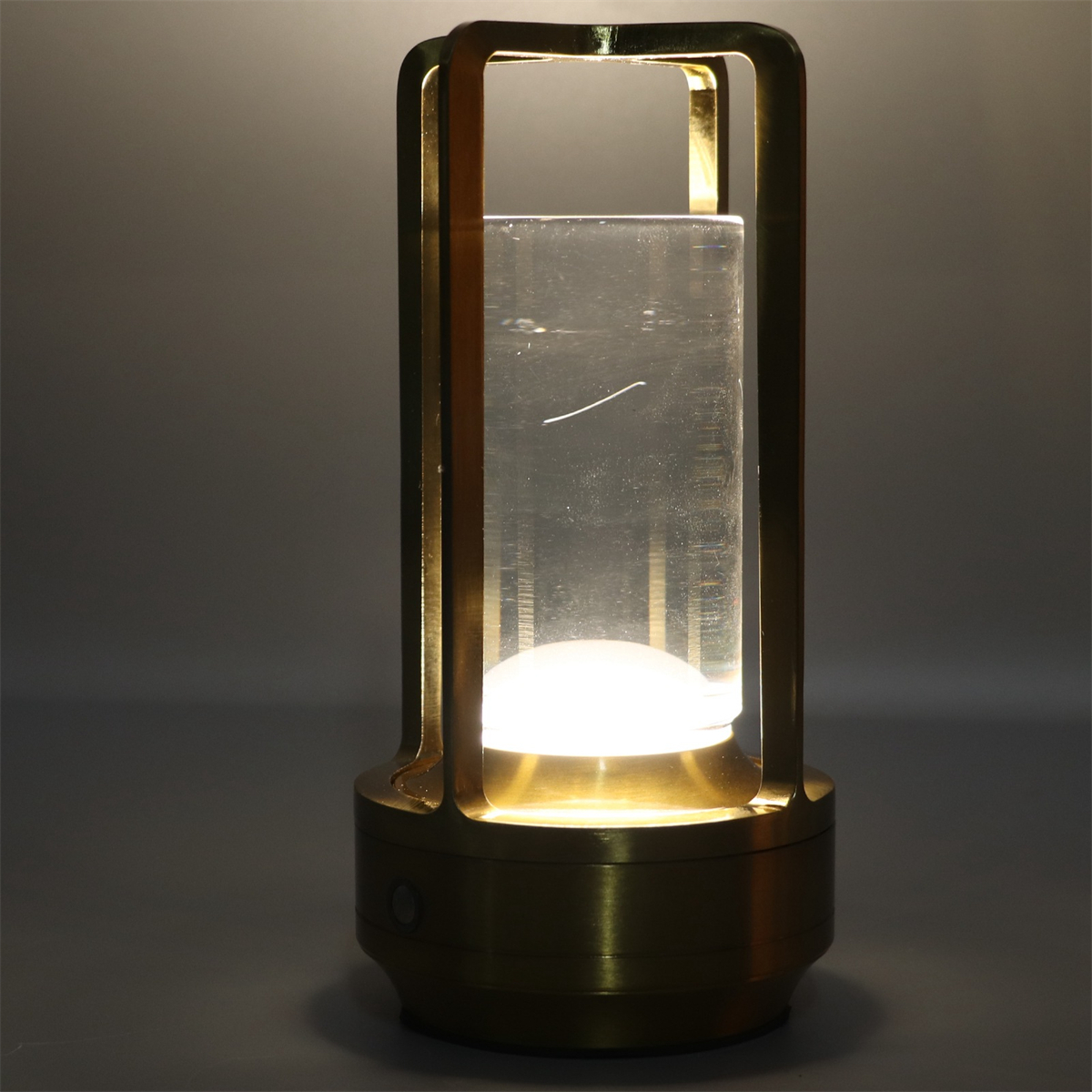 Eine goldene Laternenlampe mit einem zylindrischen Glaszentrum, das ein Licht beleuchtet. Der Hintergrund ist grau.