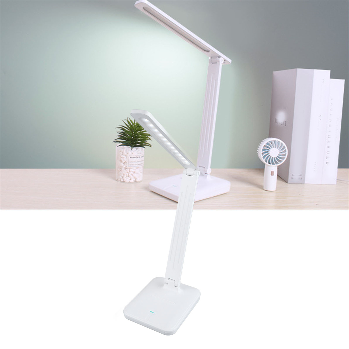 Eine weiße Schreibtischlampe mit zwei verstellbaren Lichtpaneelen, einer kleinen Pflanze und einem Ventilator.