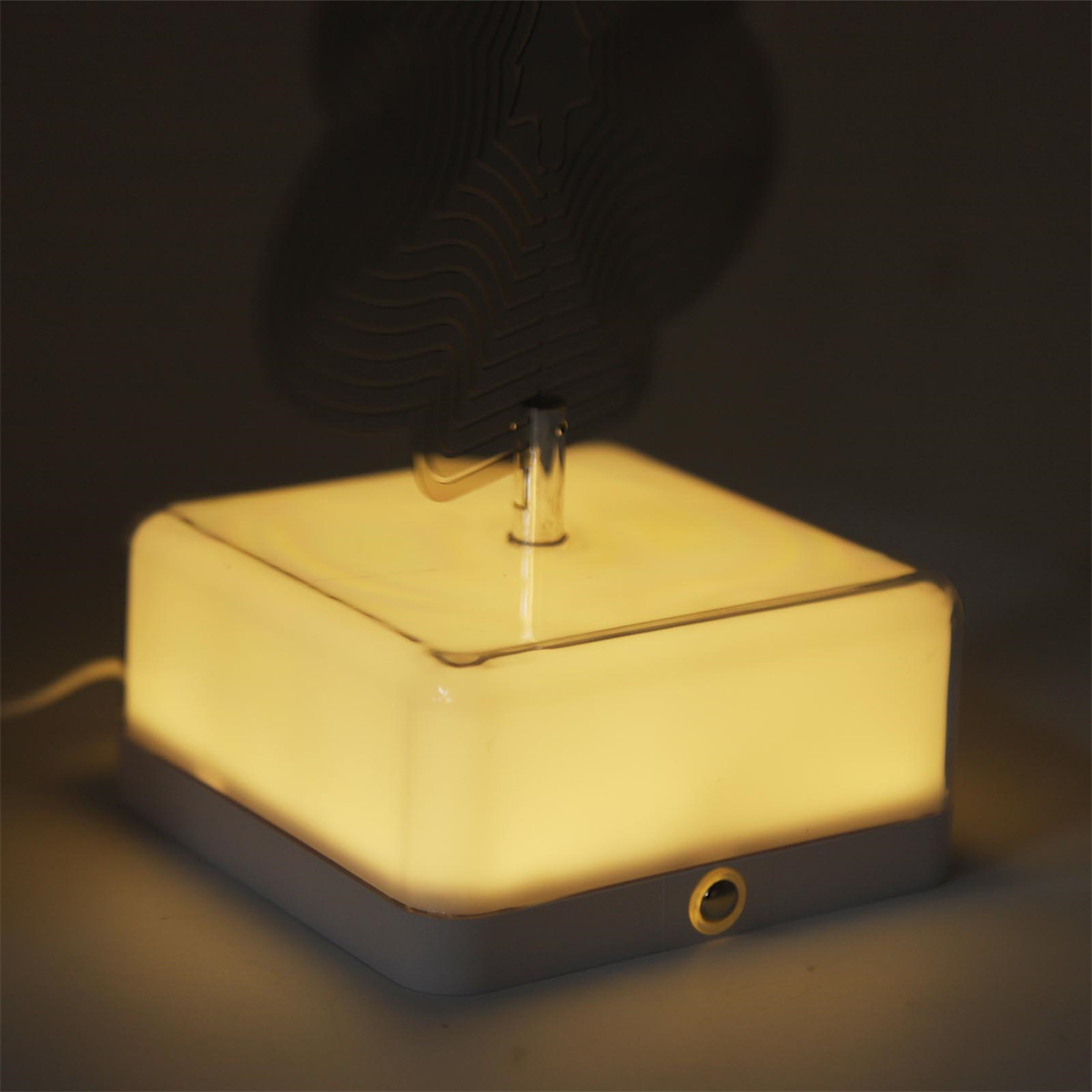 Eine beleuchtete quadratische Lampe mit fächerförmigem Aufsatz. Die Lampe ist weiß, der Fächer dunkel und der Hintergrund ist gedämpft.