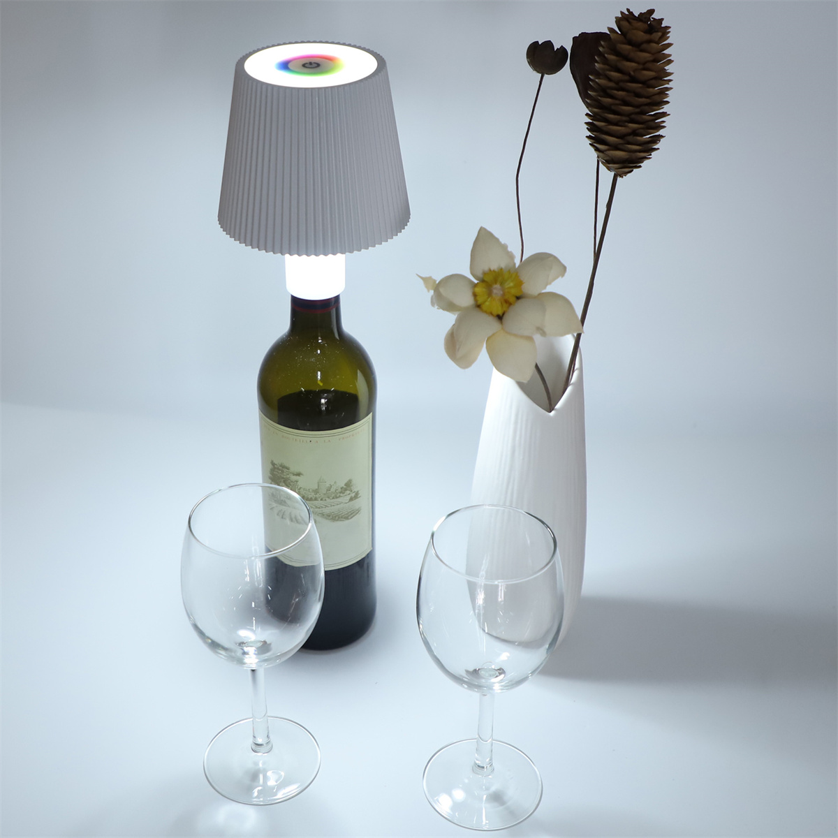 Weinflaschenlampe, Gläser und Vase mit Blume auf weißer Oberfläche.