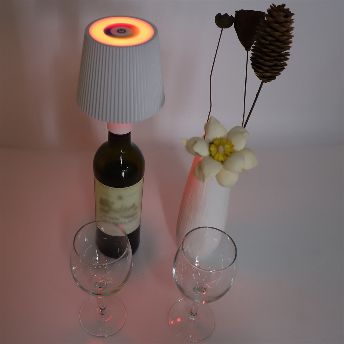 Eine Lampe auf einer Weinflasche, zwei Weingläser und eine Vase mit Blumen auf einem weißen Tisch.