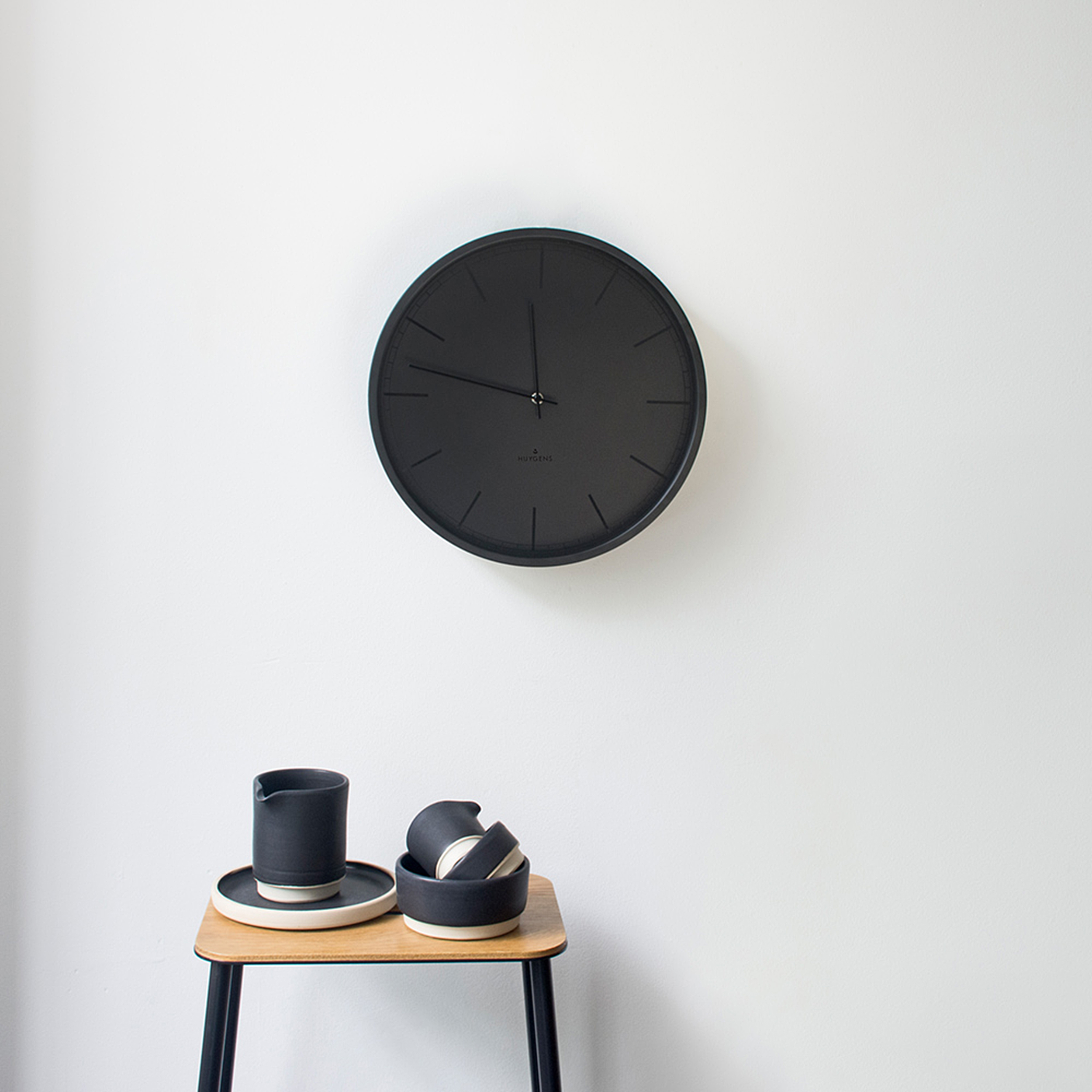 Eine schwarze Uhr an einer weißen Wand. Unter der Uhr befindet sich ein kleiner Tisch mit schwarzen Keramikartikeln.