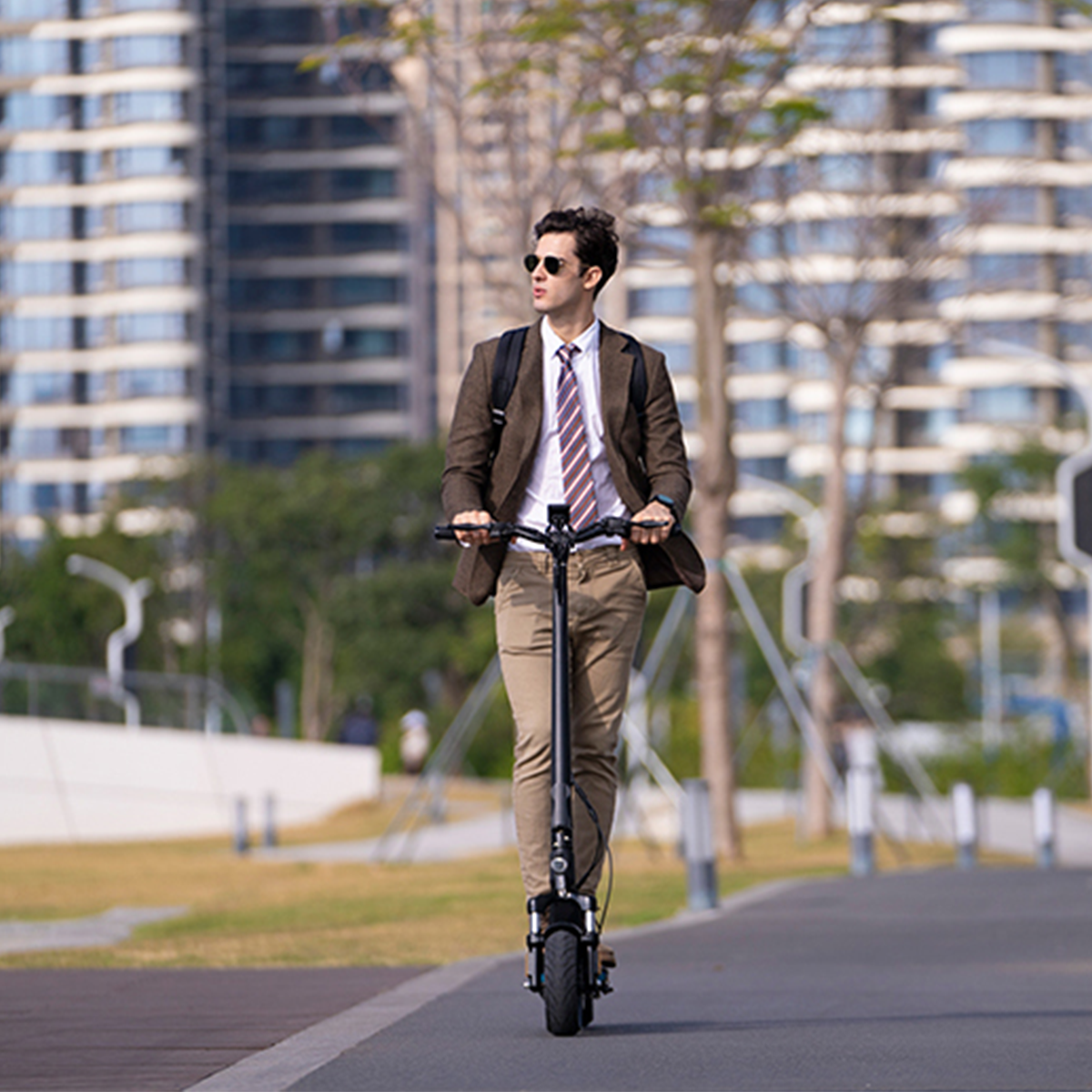Mężczyzna w garniturze jedzie na hulajnodze elektrycznej na ścieżce z tłem miasta.