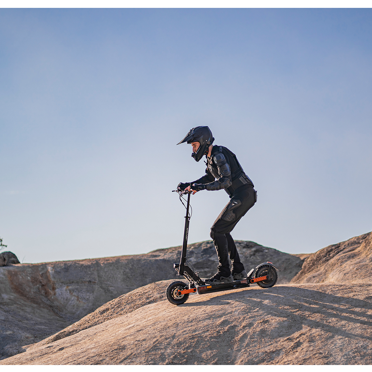 Osoba w sprzęcie ochronnym jedzie hulajnogą elektryczną po skalistym terenie pod błękitnym niebem.