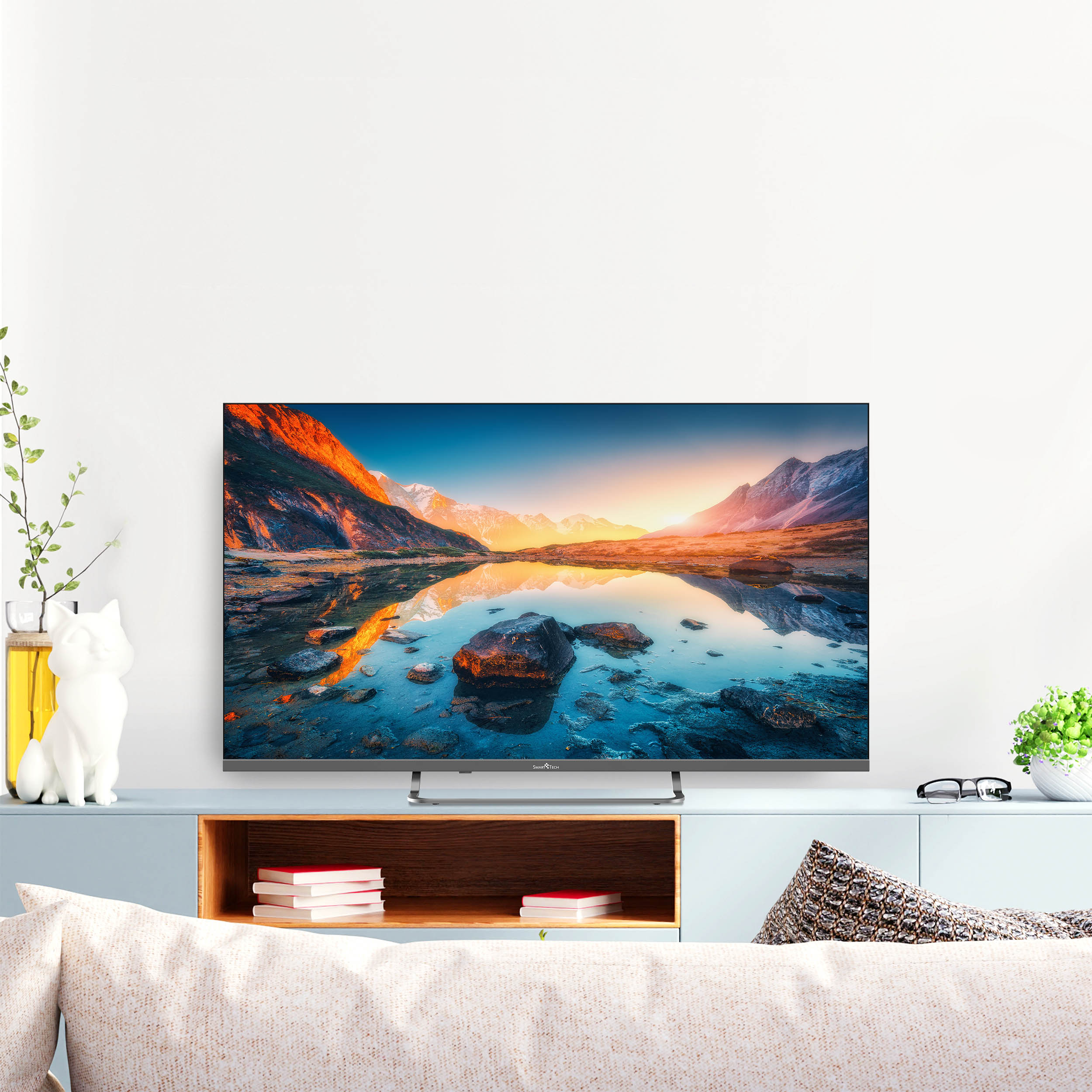 Ekran telewizora z krajobrazem górskim, na jasnoniebieskiej szafce. Biała statua kota po lewej.