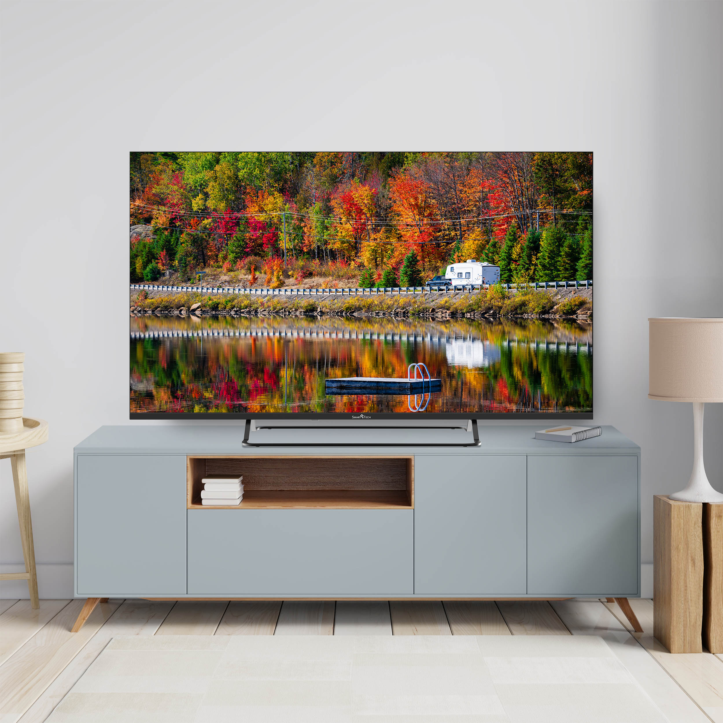 Telewizor wyświetlający kolorowy krajobraz odbijający się w wodzie. Szafka poniżej. Jesienne drzewa i pomost.