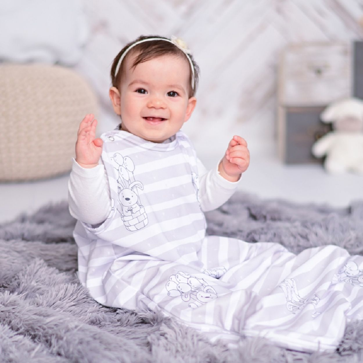 Lachendes Baby in einem Schlafsack, auf einem grauen Teppich. Ein weißes Stirnband und Hintergrund sind sichtbar.