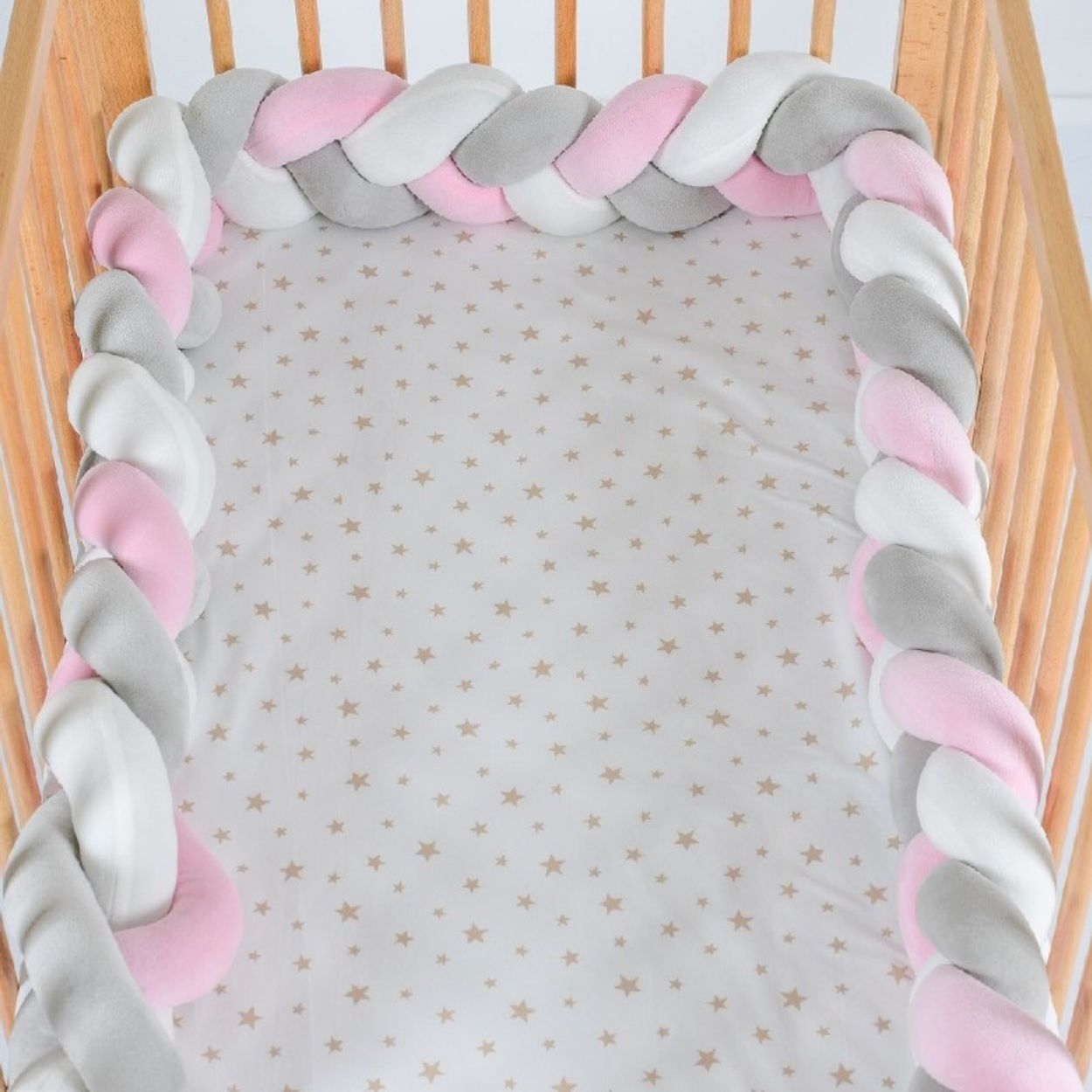 Ein Babybett mit einem geflochtenen Stoßfänger in Weiß, Rosa und Grau. Das Laken hat Sternmuster.