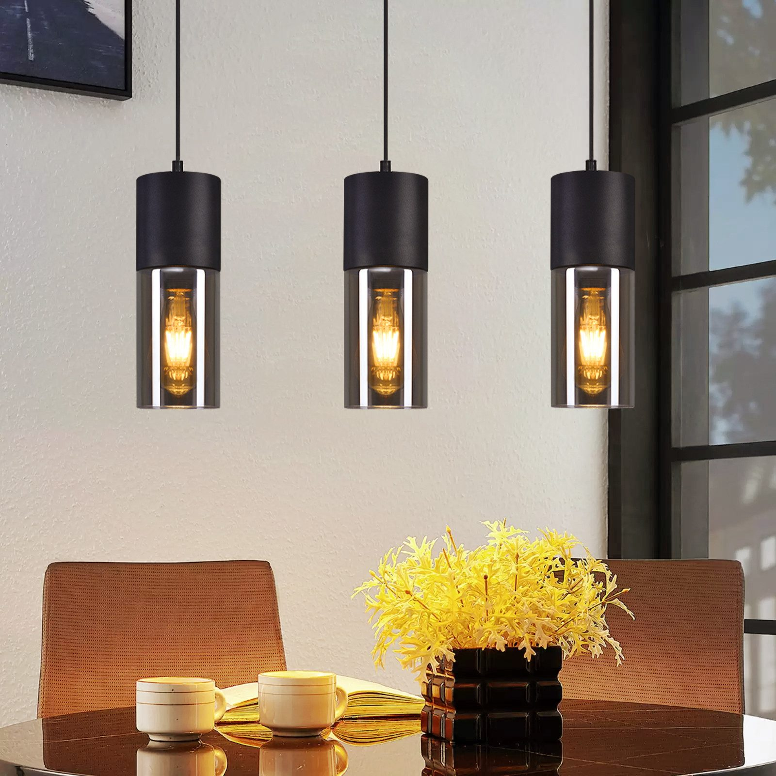 Tre lampade cilindriche nere sospese con lampadina, sopra un tavolo con fiori e tazze.