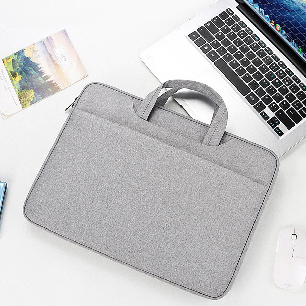 Graue Laptoptasche mit Griff, auf weißem Schreibtisch, Laptop, Maus und Notizblock sichtbar.