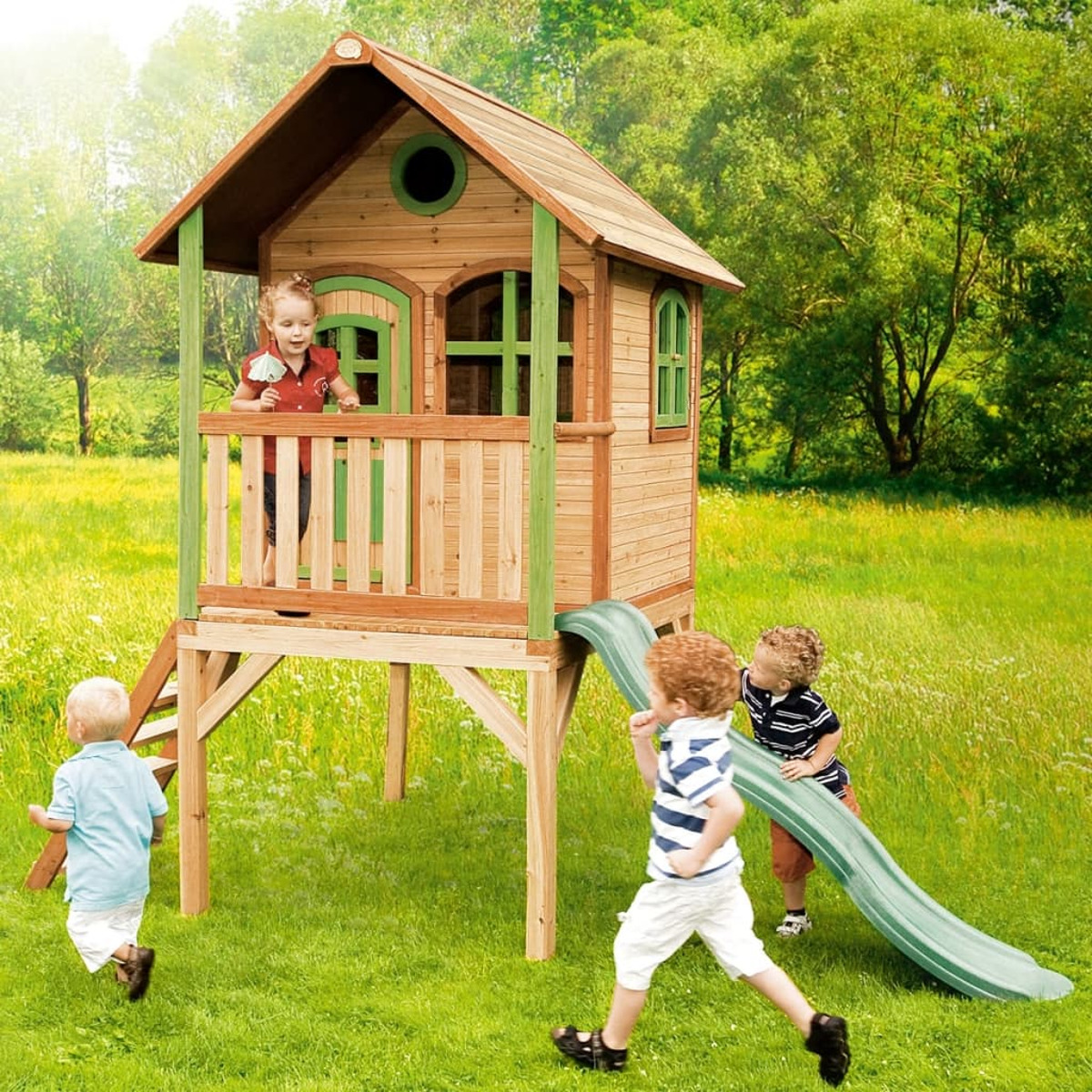 Kinder spielen in einem Holzspielhaus mit Rutsche. Grünes Gras und Bäume sind im Hintergrund.