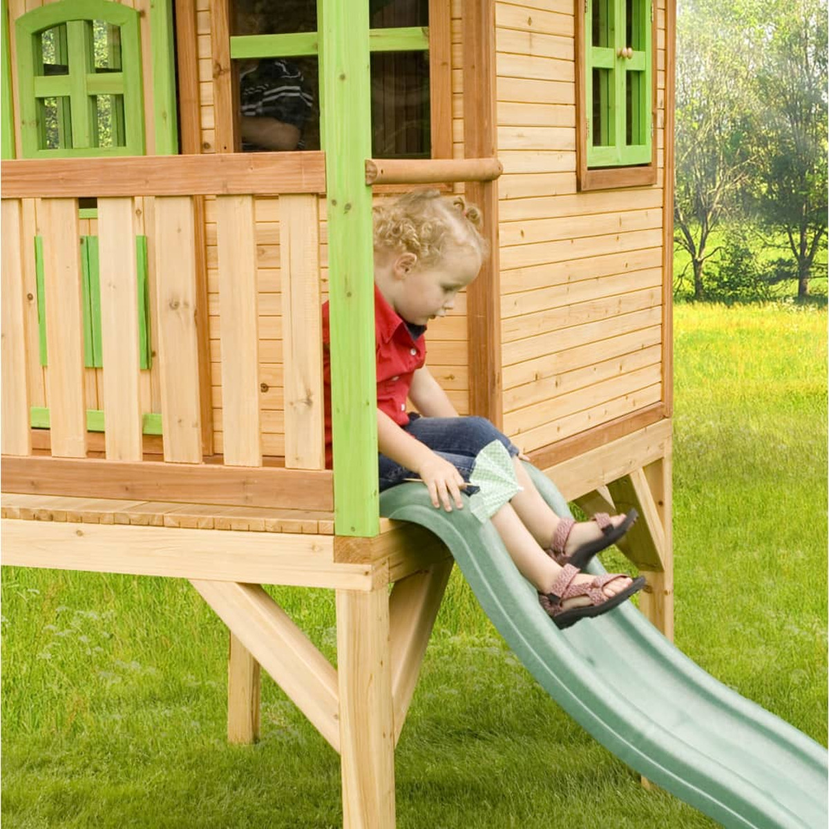 Ein Kind rutscht von einem Holzspielhaus eine grüne Rutsche hinunter. Darunter grünes Gras.